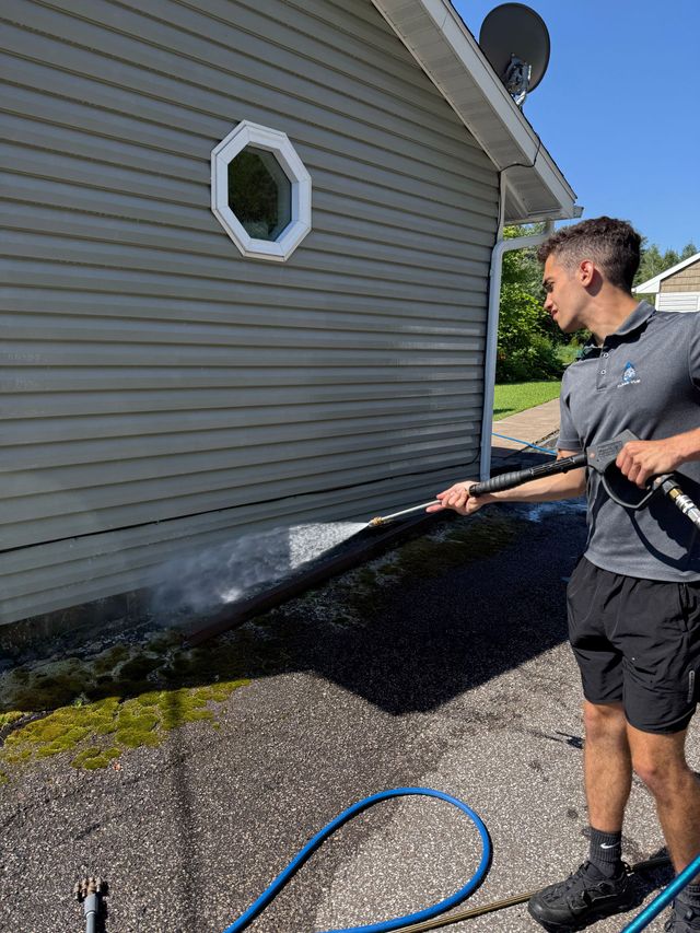Man spraying water on a gray house siding near the foundation with a hose outdoors