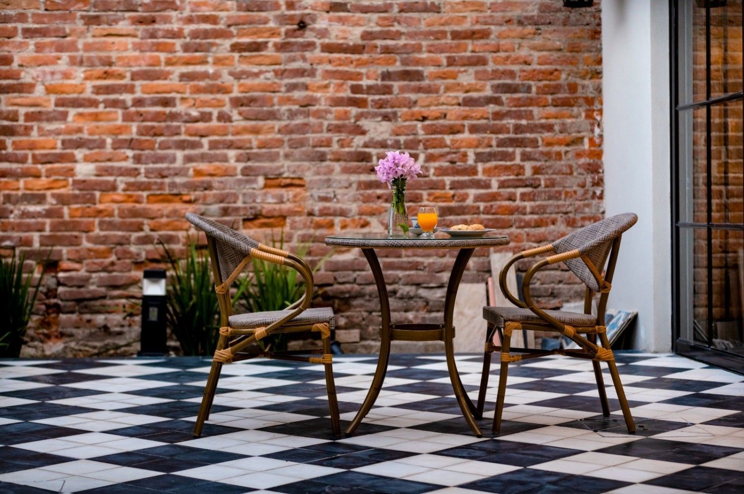 Sillas de mimbre y una mesa en un patio a cuadros contra una pared de ladrillos, con flores y un vaso de jugo.