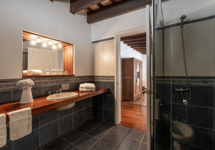 Baño con suelo de baldosas oscuras, lavabo de madera y ducha. La puerta da a otra habitación con suelo de madera.