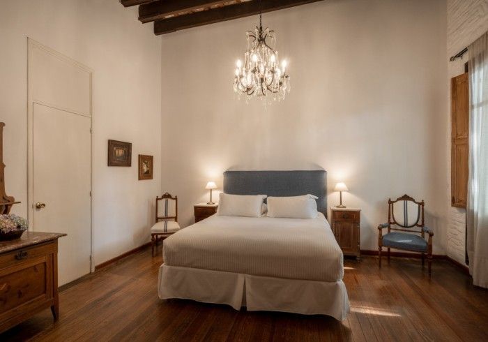 Dormitorio con cama, mesitas de noche, lámpara de araña, suelo de madera, sillas y paredes blancas.