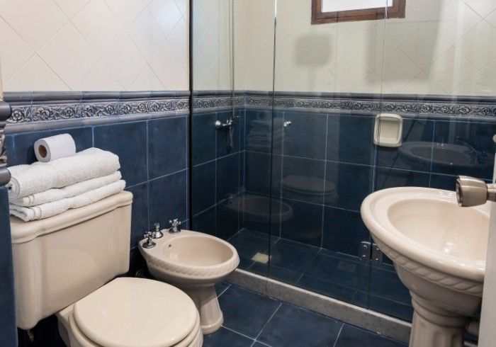 Baño con azulejos azules, inodoro, bidé, lavabo y ducha. Toallas blancas en el inodoro.