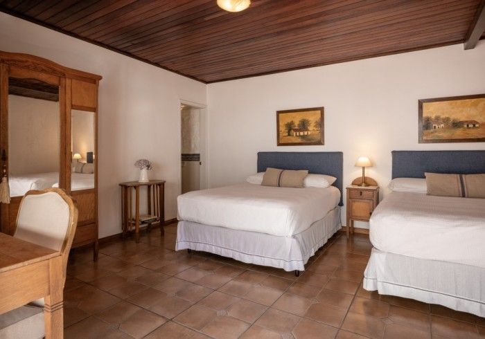 Dormitorio con dos camas, cómoda, escritorio y mesita de noche. Paredes beige y blancas, techo y suelo de madera.