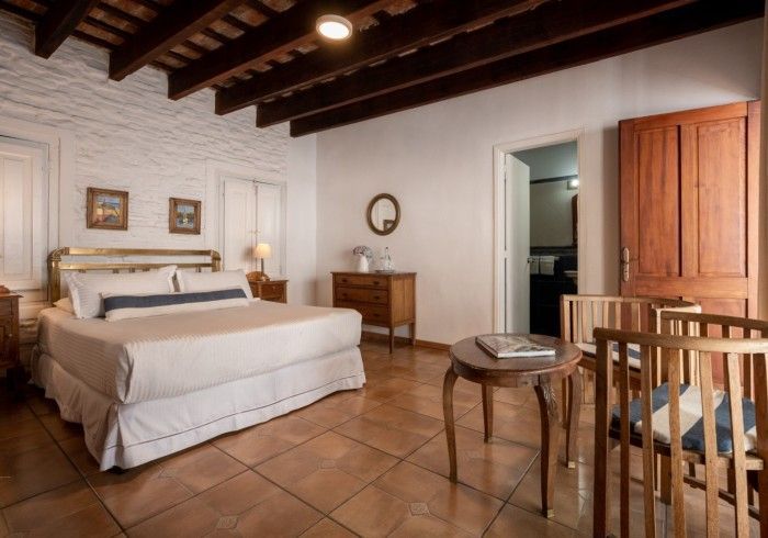 Dormitorio con cama, cómoda, mesa y sillas. Paredes beige, techo y puerta de madera, suelo de baldosas.