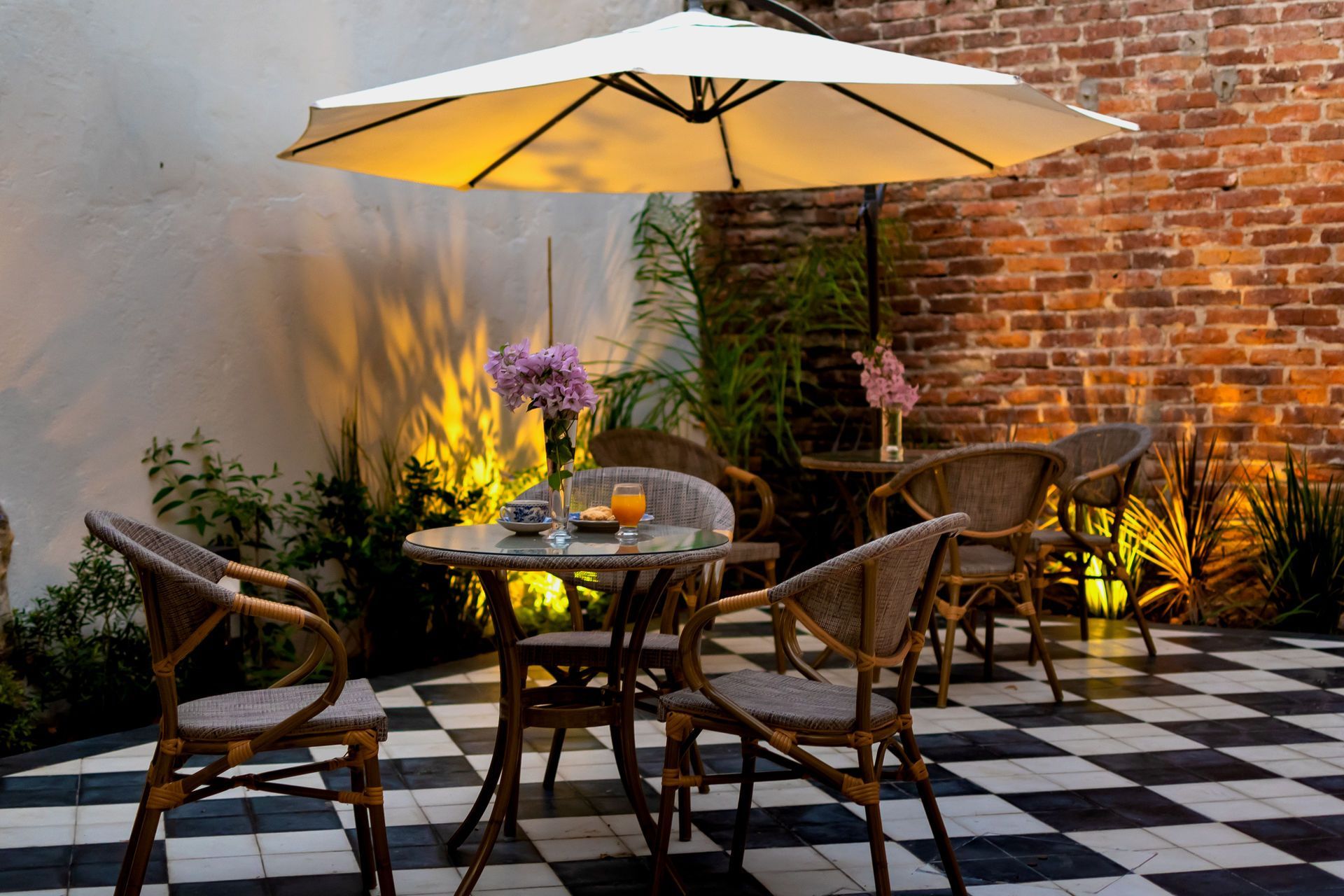Patio con mesa, sillas, sombrilla, flores, pared de ladrillo y suelo a cuadros blancos y negros.