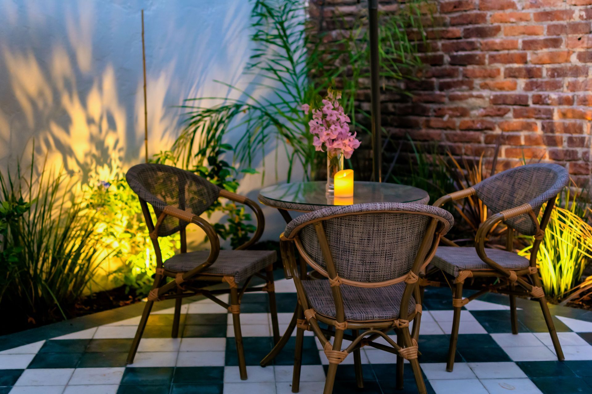 Conjunto de comedor de patio: mesa redonda, cuatro sillas de mimbre, plantas, pared de ladrillo e iluminación suave.