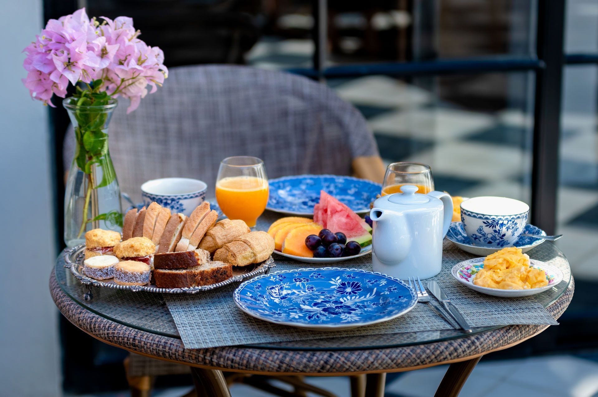 Mesa preparada para el desayuno: bollería, fruta, zumo, tetera, platos florales y jarrón con flores rosas.