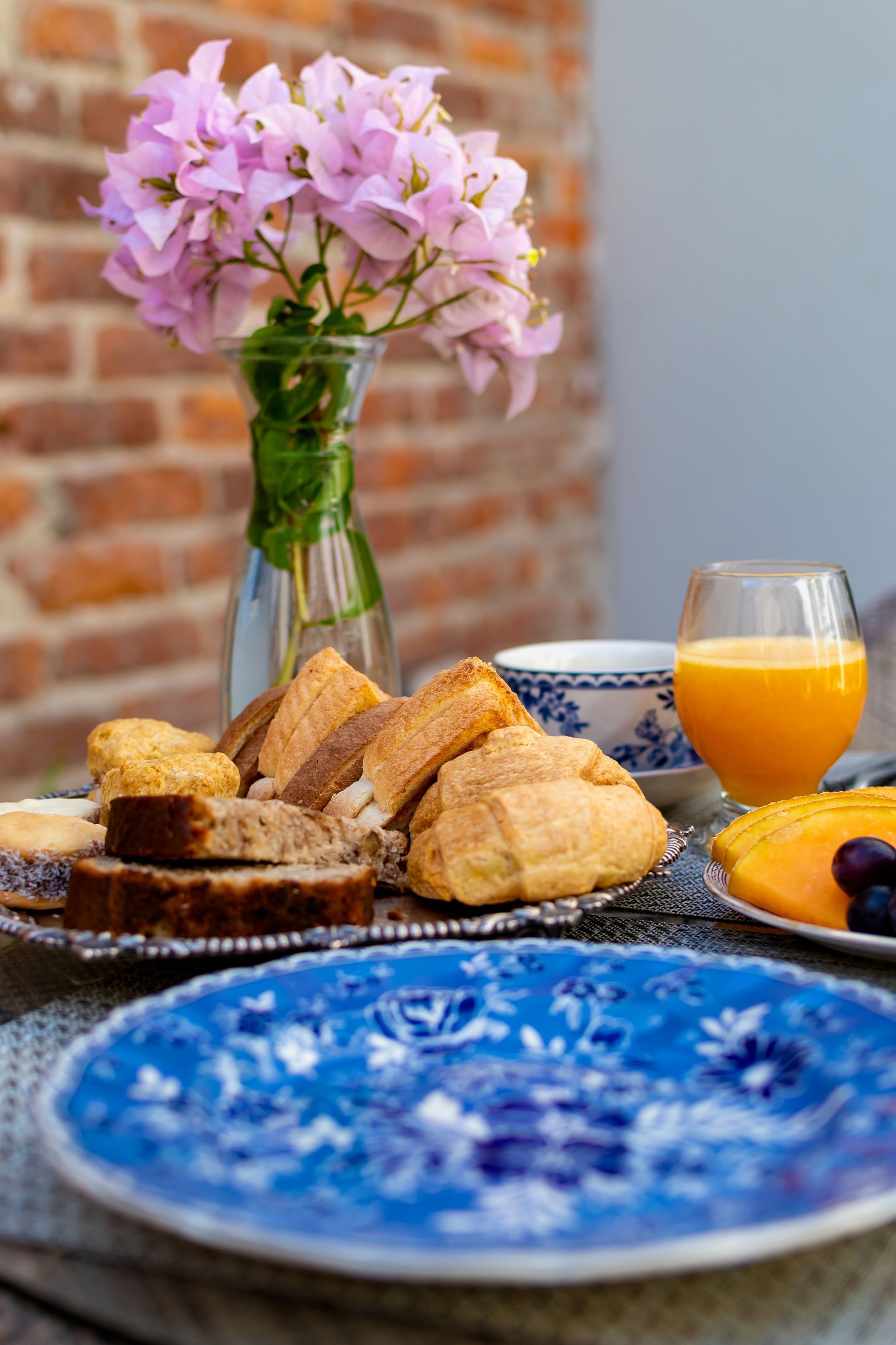 Mesa de desayuno con pan, flores y jugo, contra una pared de ladrillos.