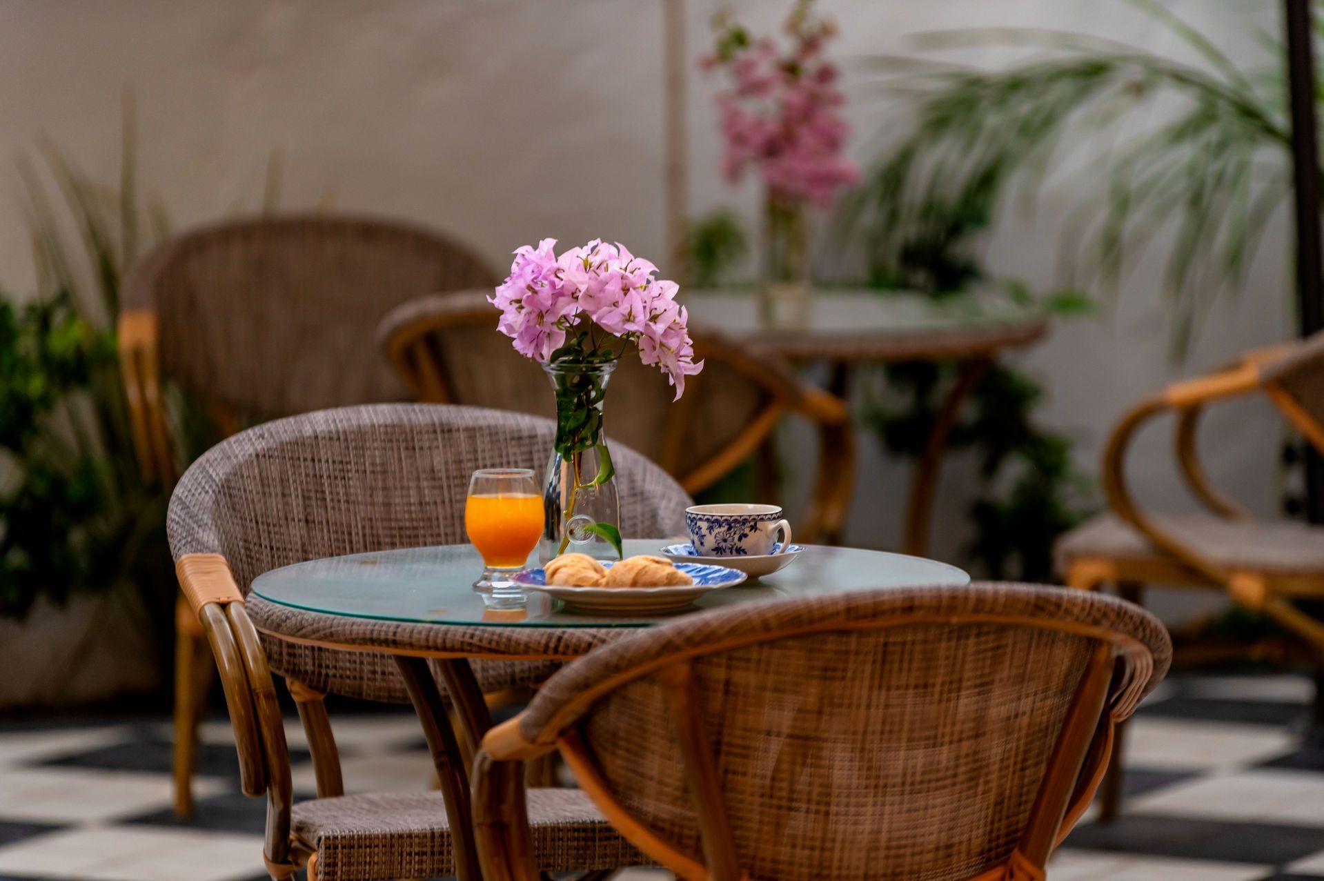 Mesa de mimbre con pasteles, jugo de naranja y flores, en un ambiente de patio con piso a cuadros.