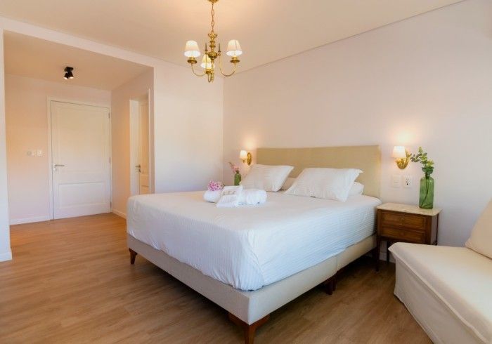 Dormitorio con cama king size, mesita de noche y sofá pequeño. Paredes blancas y suelo de madera.