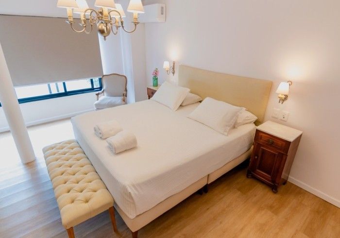 Dormitorio con cama beige, banco y ventana. Cerca hay una mesita de noche y una silla.