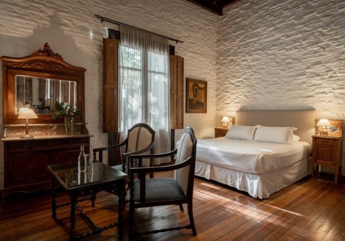 Elegante dormitorio con pared de ladrillo blanco, cama doble, muebles antiguos y gran ventanal.