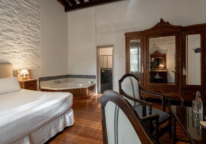 Habitación de hotel con cama, jacuzzi, armario antiguo, sillas y puerta de baño. Suelos de madera.