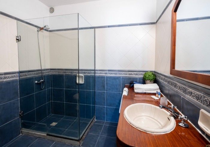 Baño con azulejos azules, ducha de cristal, tocador de madera y lavabo blanco.