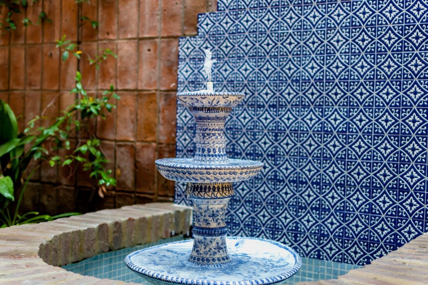 Fuente con diseño de azulejos azules y blancos, con agua fluyendo. Fondo de pared de azulejos, ladrillos y vegetación.