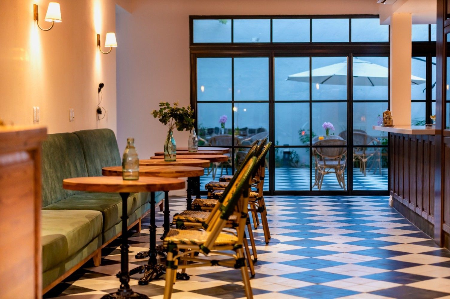 Interior de restaurante con un stand verde, mesas y suelo a cuadros, con vistas a un patio con sombrilla.