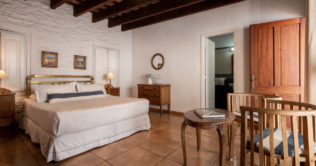 Dormitorio con cama, mesitas de noche, cómoda, mesa pequeña y sillas; puerta de acceso al baño.