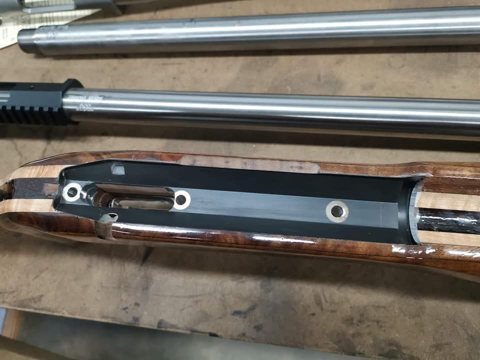 Long silver firearm barrel and black part resting in a camo-patterned gun case on a workbench