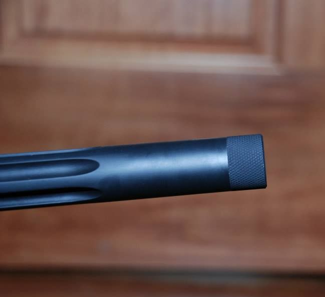 Close-up of a black firearm barrel with a textured muzzle brake against a wooden background