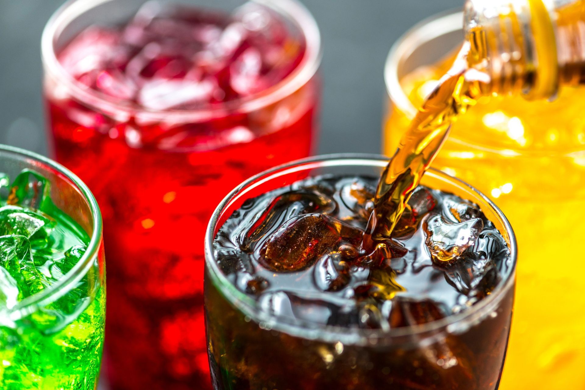 Four glasses of colorful soda with ice; brown soda being poured into one.