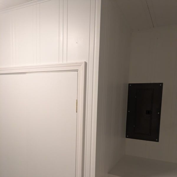 White hallway wall with a closed door and a recessed black electrical panel on the right.