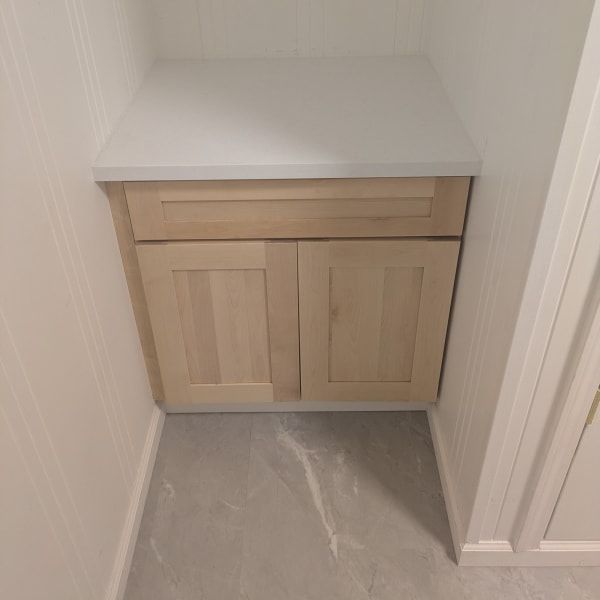 Light wood bathroom vanity cabinet with white countertop in a narrow alcove, on gray floor.