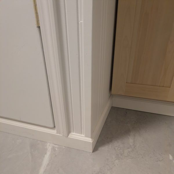 Corner of a kitchen with white trim, gray floor, and a light wood cabinet panel