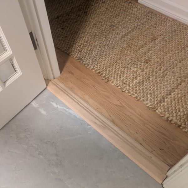 Threshold between gray floor and wood hallway carpet, with an open white door.