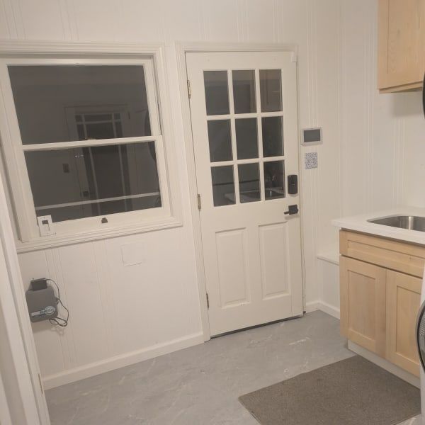 Small kitchen with white door and window, light wood cabinets, and a gray floor mat.