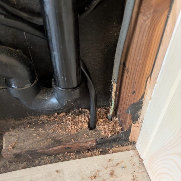 Damaged, rotting wood around black pipes beside a wall and floor trim in a cramped corner