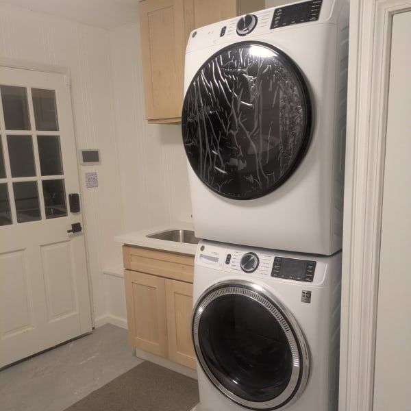 Stacked white washer and dryer in a laundry room with light wood cabinets and a sink.