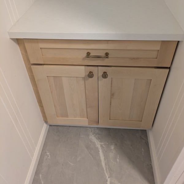 Light wood bathroom vanity cabinet with drawers and double doors in a narrow alcove, over gray tile floor