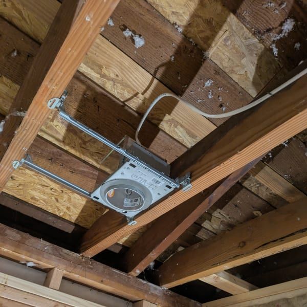Ceiling-mounted speaker in exposed attic framing with wooden joists and insulation visible.