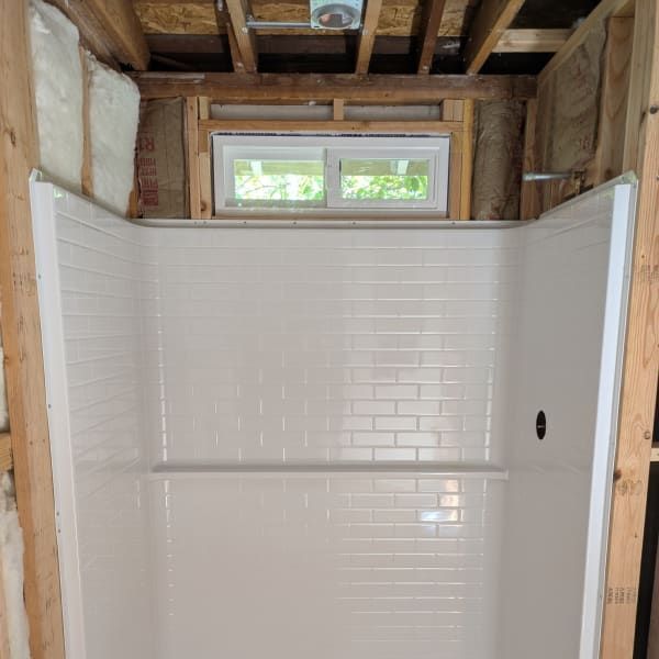 Tiled shower installed in an unfinished bathroom with exposed framing and a small window above.