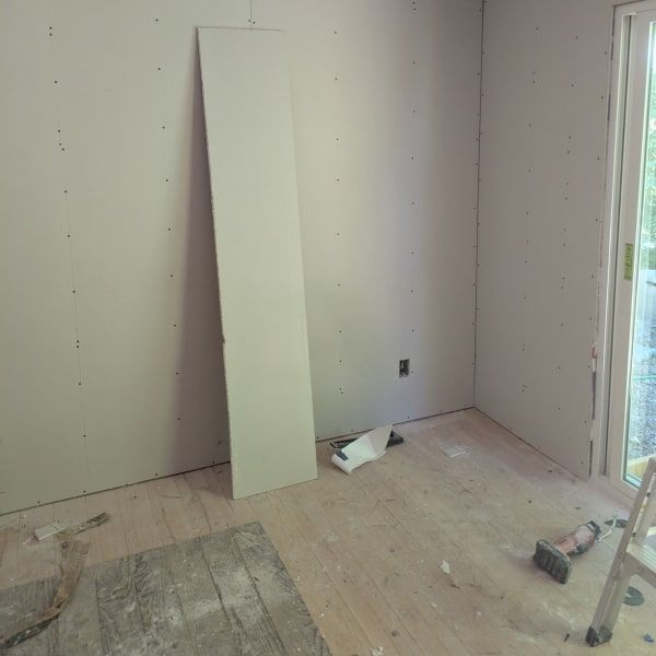 Unfinished room with drywall, leaning board, tools on floor, and an open door to outside.
