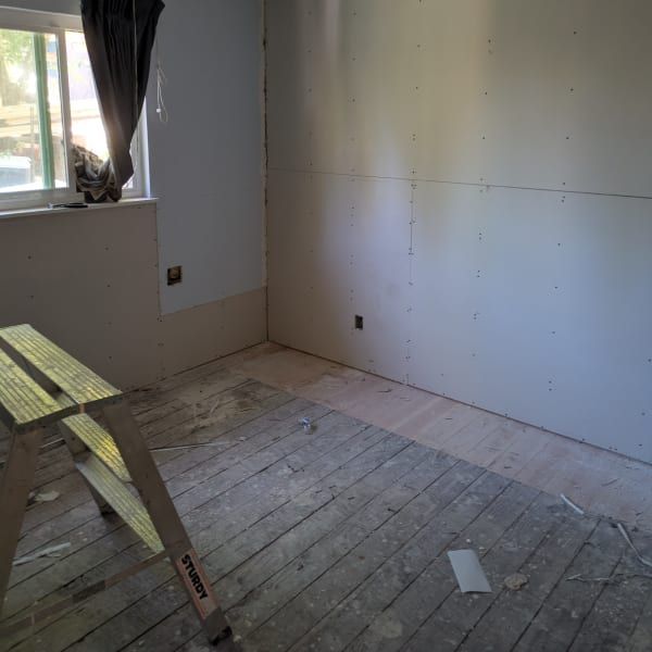 Empty room under renovation with bare drywall, unfinished floor, a window, and a saw on sawhorses.