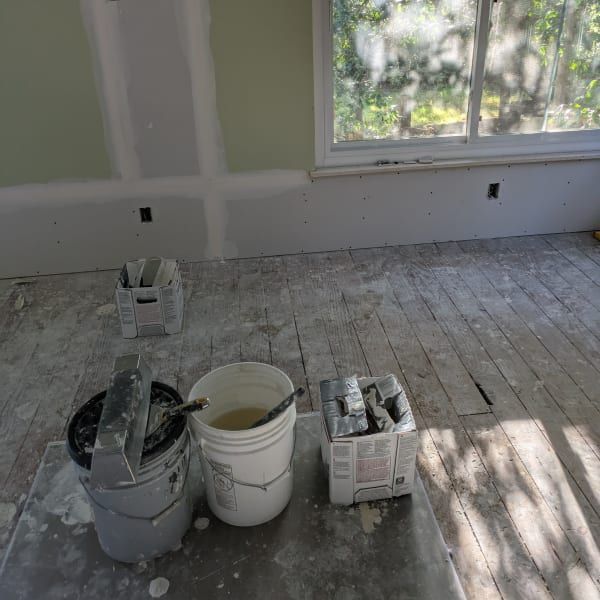 Paint buckets and supplies on a dusty floor in a room being painted, with a window letting in sunlight