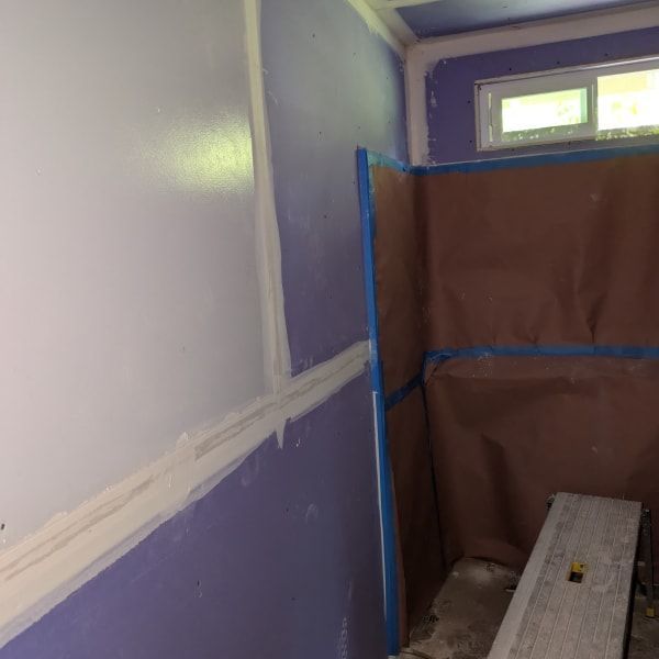 Taped-off bathroom walls being painted white and purple, with a covered window and ladder in the corner