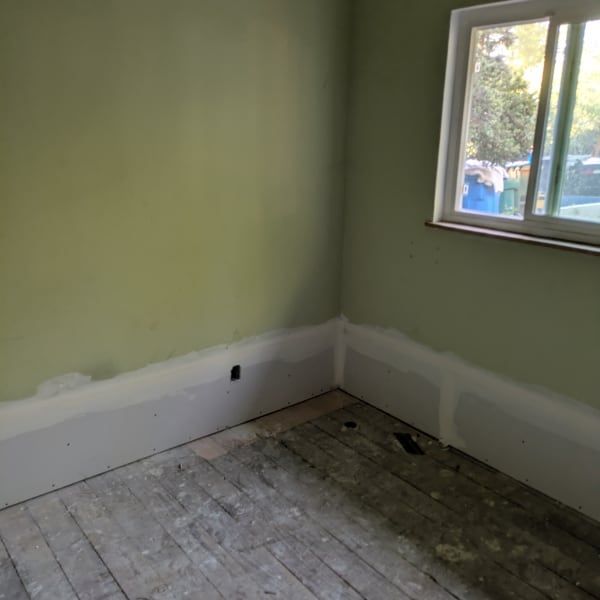 Empty room with green walls, white baseboards, unfinished floor, and a window on the right.