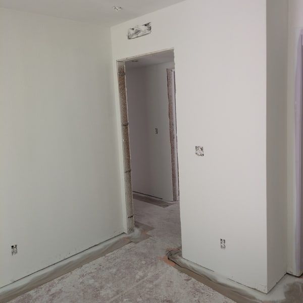 Empty white room under renovation with bare walls, unfinished floor, and a doorway into a hall