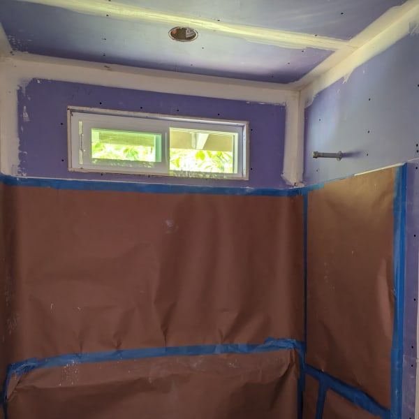 Small room under renovation with purple walls, brown protective coverings, blue tape, and a small window.