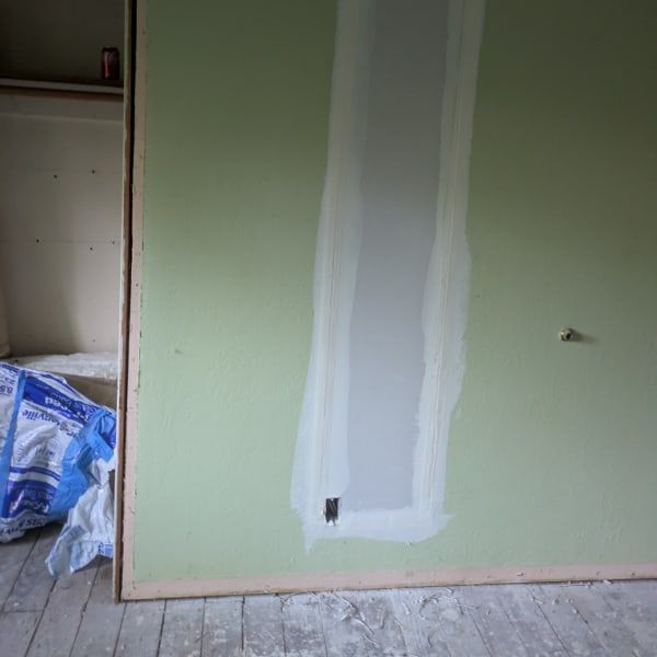 Partially painted green wall with a white vertical patch and outlet hole in an unfinished room