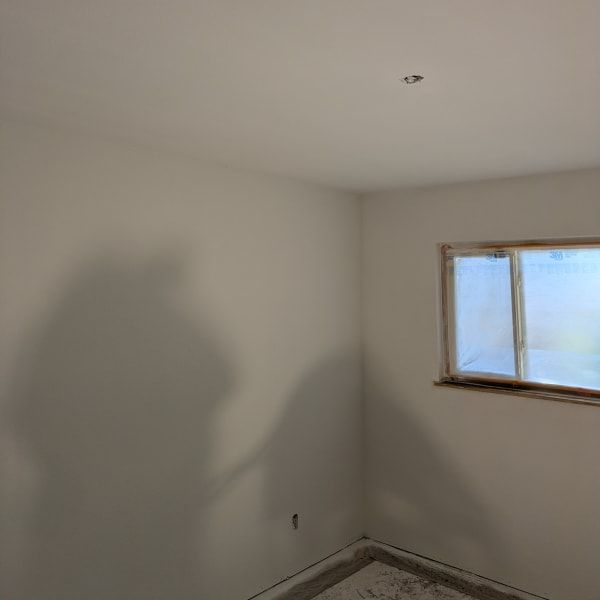 Empty white room with a window, unfinished baseboards, and a ceiling light fixture.