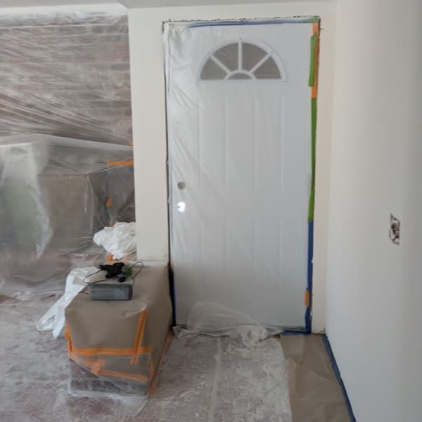 White front door in a room under renovation, with plastic-covered furniture and painter’s tape nearby.