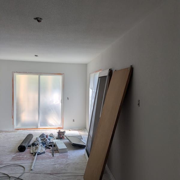 Renovated living room with white walls, bare floor, scattered tools, and large boards leaning against the wall.