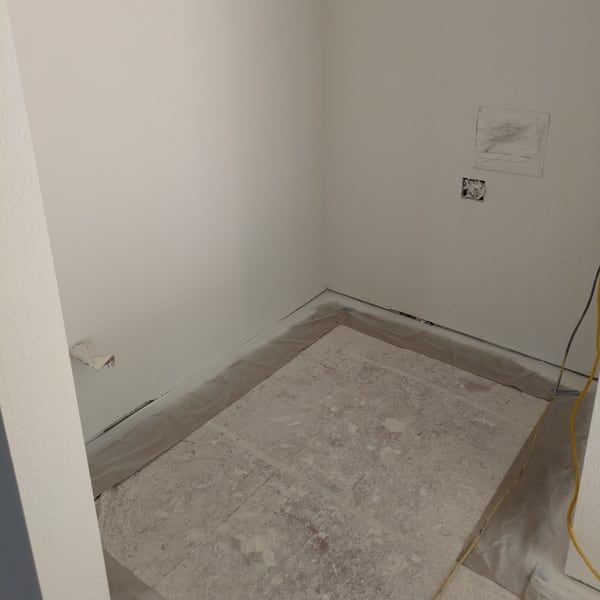Empty white room corner with bare walls, floor covering, and exposed wiring during renovation
