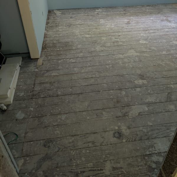 Empty room with worn, unfinished wooden floorboards and pale blue walls.