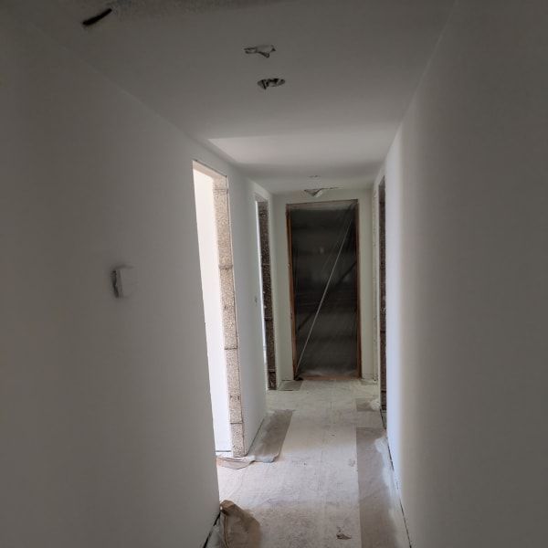 Narrow white hallway under construction with unfinished walls, exposed doorways, and debris on the floor