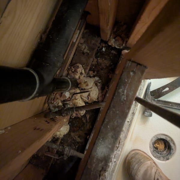 Cluttered floor cavity with exposed pipes, wood framing, debris, and a boot near a drain opening