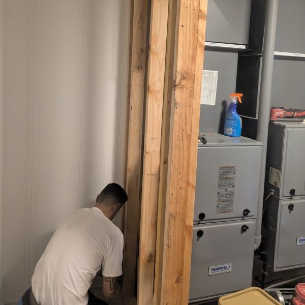 Worker crouching by HVAC units in an unfinished utility room with exposed wood framing.