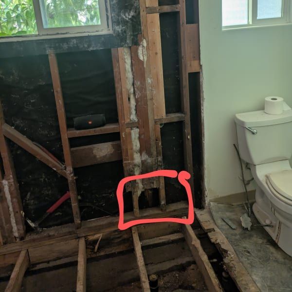 Damaged bathroom with exposed wall studs and floor joists beside a toilet; red mark highlights a floor opening